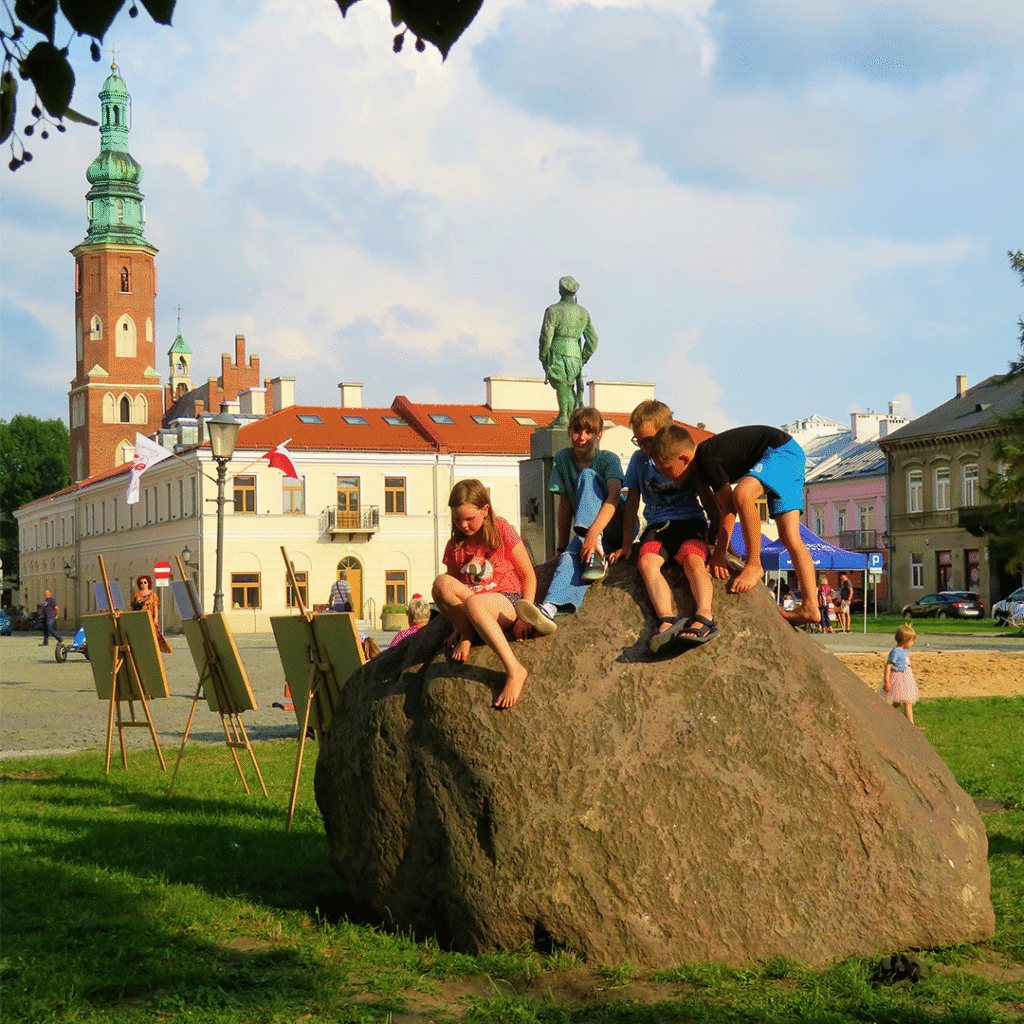 Grupa dzieci bawi się na dużym kamieniu na radomskim Rynku. W tle znajduje się pomnik, zabytkowy budynek Kamienicy Deskurów oraz wieża fary, kamienice oraz sztalugi. Niebo jest częściowo zachmurzone, a na trawie widoczne są inne osoby.