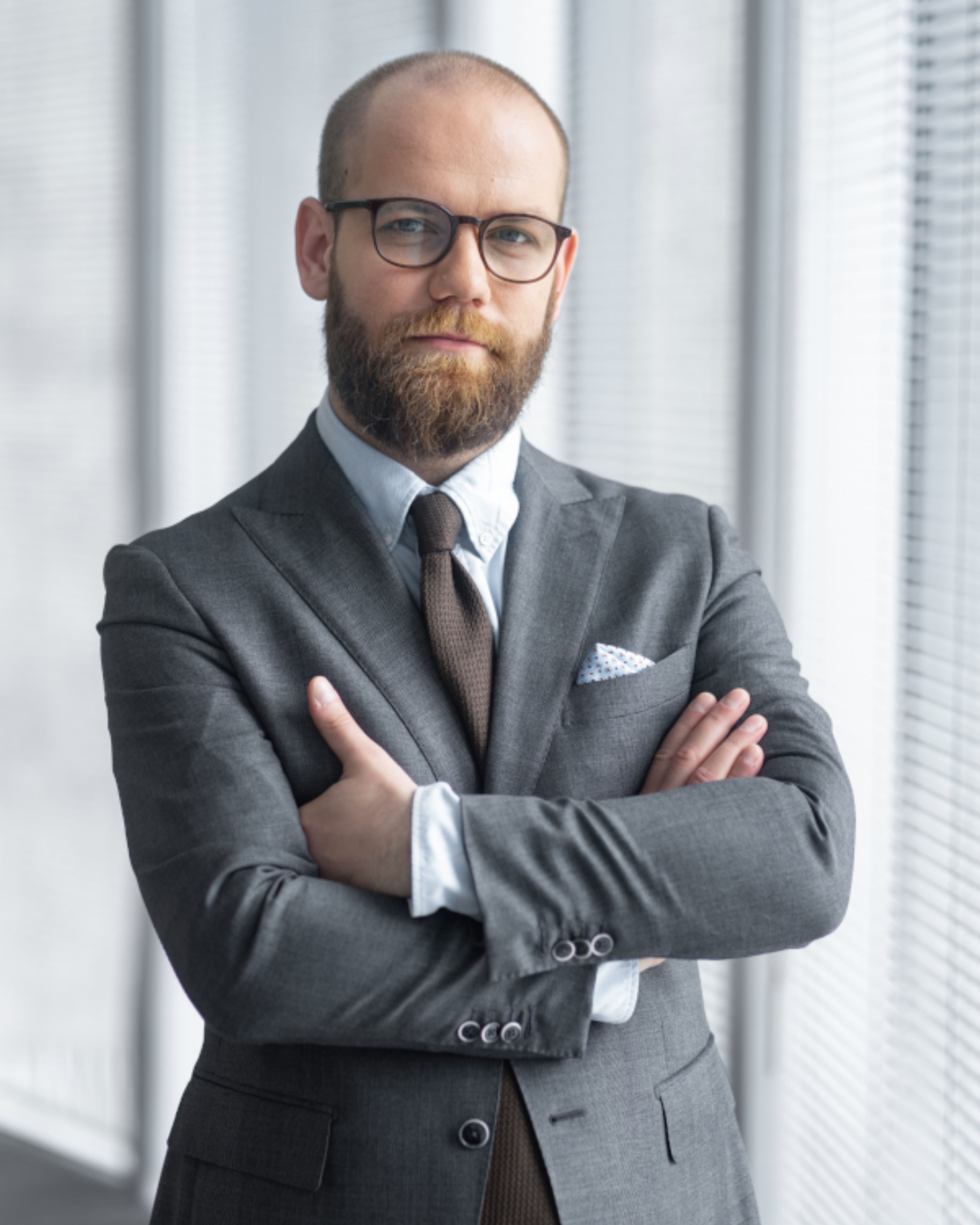 „Mężczyzna z brodą i okularami w eleganckim szarym garniturze, błękitnej koszuli i brązowym krawacie, stoi z założonymi rękami na tle dużych okien z żaluzjami. Patrzy poważnie w stronę aparatu.”