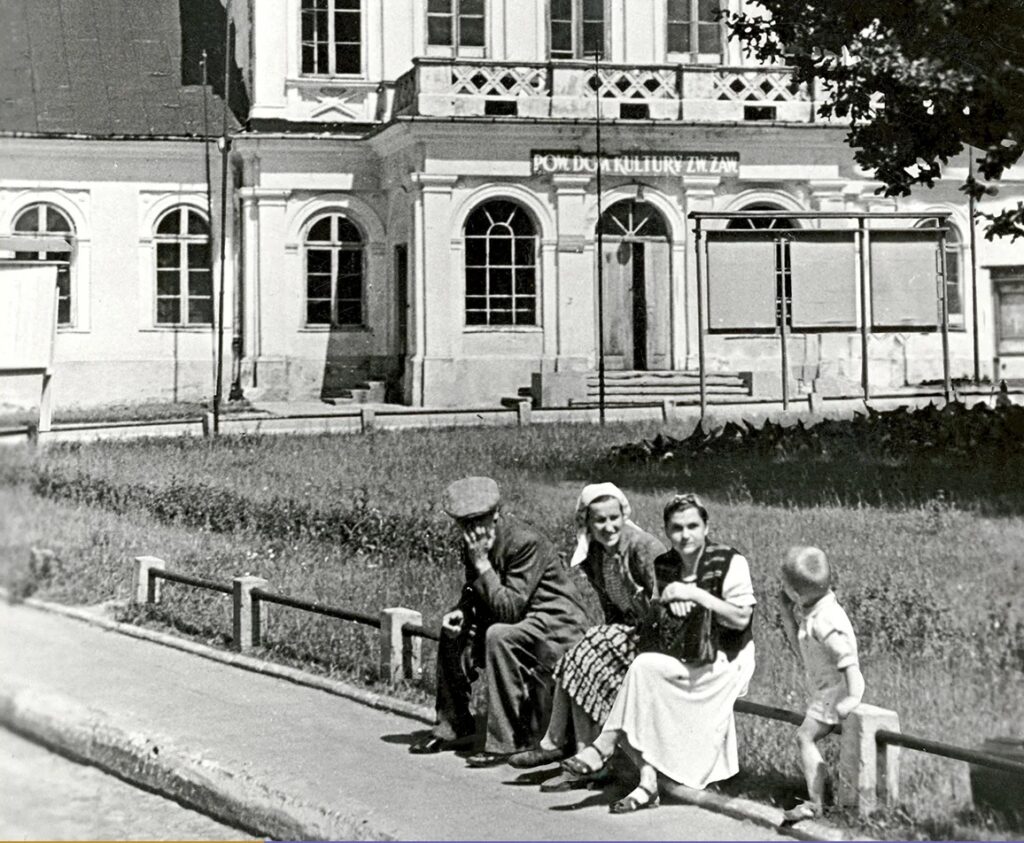 Czarno-białe zdjęcie historyczne przedstawiające grupę osób (mężczyznę, dwie kobiety i dziecko) siedzących na niskim ogrodzeniu przy chodniku. W tle widoczny jest jasny, klasycystyczny budynek z napisem „POW. DOM KULTURY ZW. ZAW.” nad wejściem.