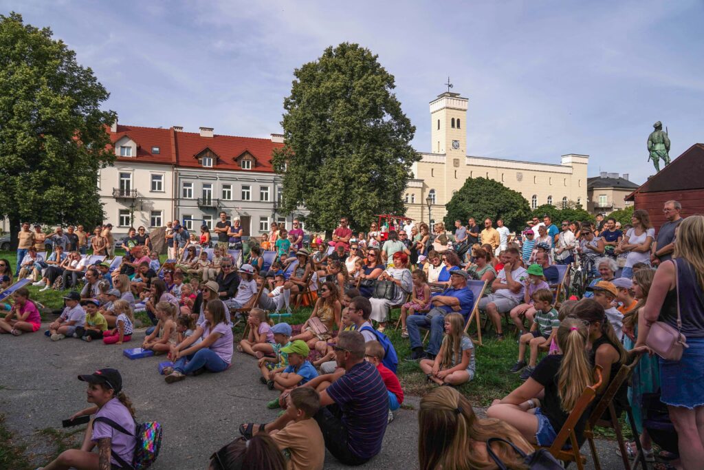 Licznie zgromadzona publiczność, składająca się z dorosłych i dzieci, siedzi na leżakach oraz na ziemi podczas plenerowego wydarzenia na rynku miejskim. W tle widoczne są kamienice, duże drzewa oraz charakterystyczny biały budynek z wieżą.