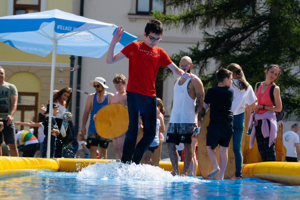Chłopak w czerwonej koszulce i ciemnych spodniach ślizga się na wodnym torze typu skimboard w przestrzeni miejskiej, wywołując duży rozbryzg wody. Wokół toru stoją obserwujący go dorośli i dzieci.