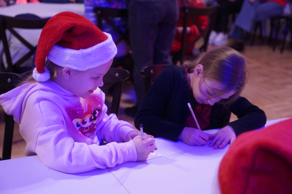 Zdjęcie przedstawia dwie dziewczynki siedzące przy białym stoliku i rysujące. Dziewczynka po lewej stronie ma na głowie czerwoną czapkę Mikołaja z białym wykończeniem i ubrana jest w jasnoróżową bluzę z kapturem, natomiast dziewczynka po prawej ma ciemny ubiór i pochyla się skupiona nad kartką, trzymając w ręku długopis lub kredkę.